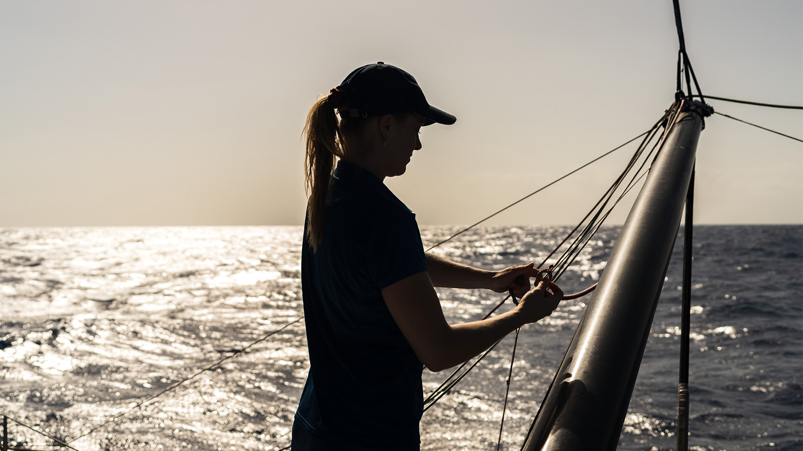 A woman on a boat, silhouetted against a bright sky, adjusts the sails while navigating on the open ocean.