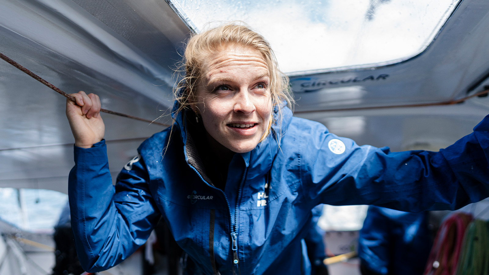 A person in a blue rain jacket is leaning out from a window or hatch on a boat, looking determined, with wet hair slightly disheveled.