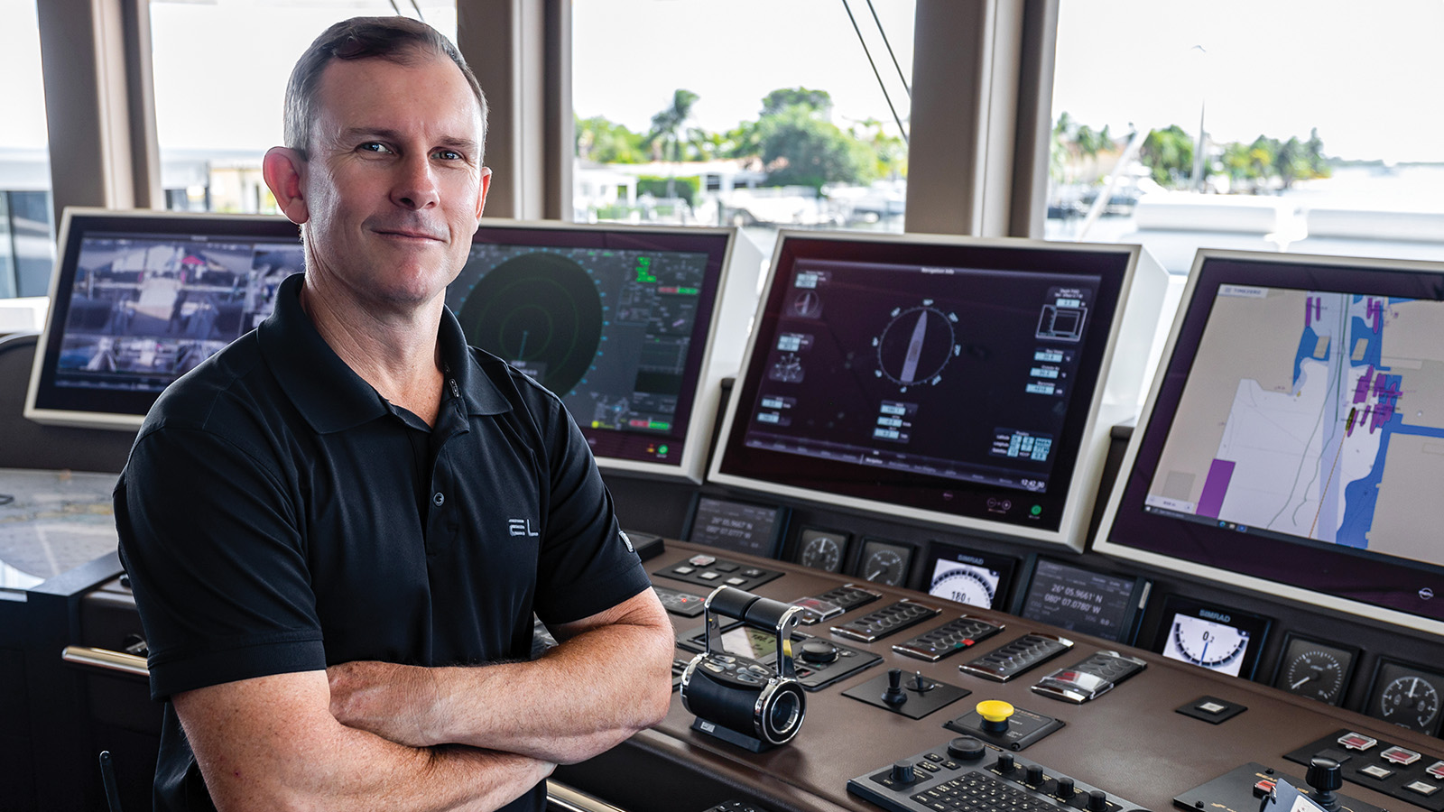 A person wearing a black polo shirt stands with arms crossed in a ship's control room, surrounded by several electronic navigation screens and equipment.