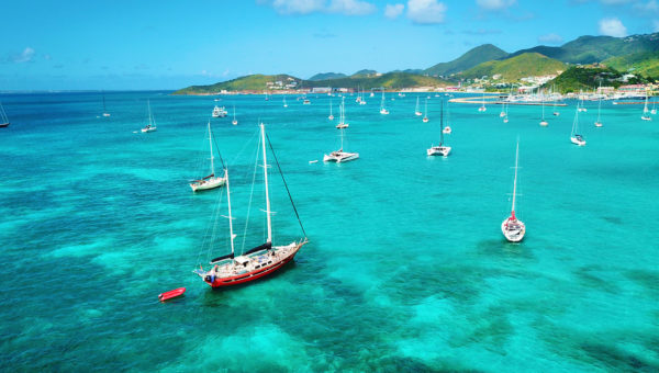 A vibrant coastal scene with numerous sailboats and catamarans anchored in clear turquoise waters. The background features lush green hills under a blue sky with scattered clouds, suggesting an idyllic and serene setting.