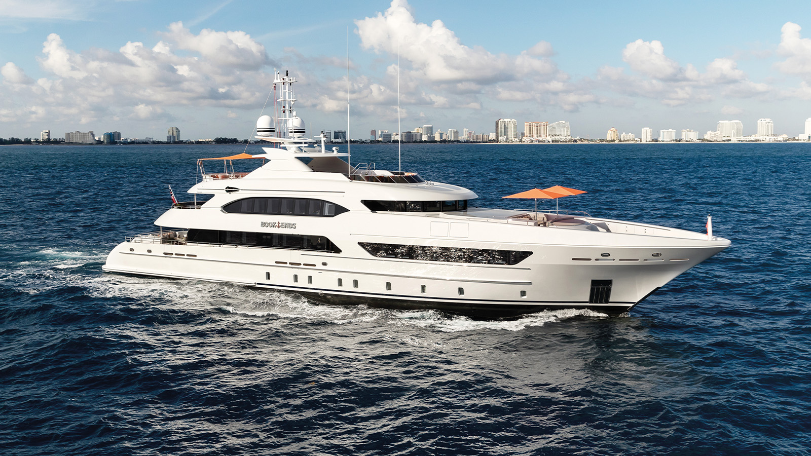 A large luxury yacht named 'Bookends' cruising on the ocean with a city skyline visible in the background.