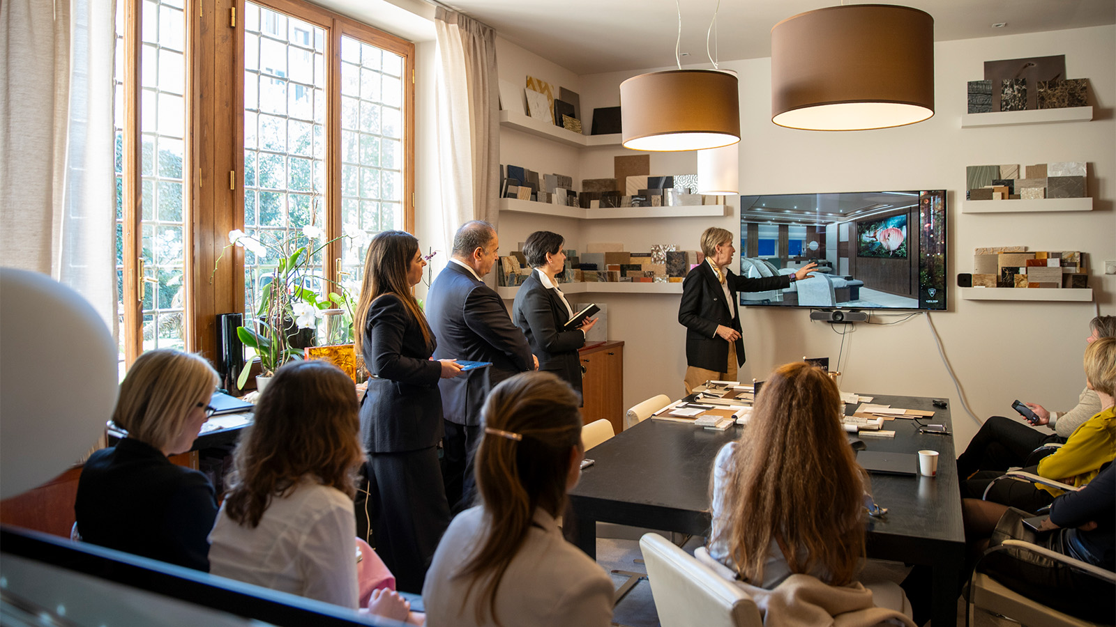 A group of people in a meeting room watching a presentation on a wall-mounted screen, with samples displayed on shelves and a large window providing natural light.