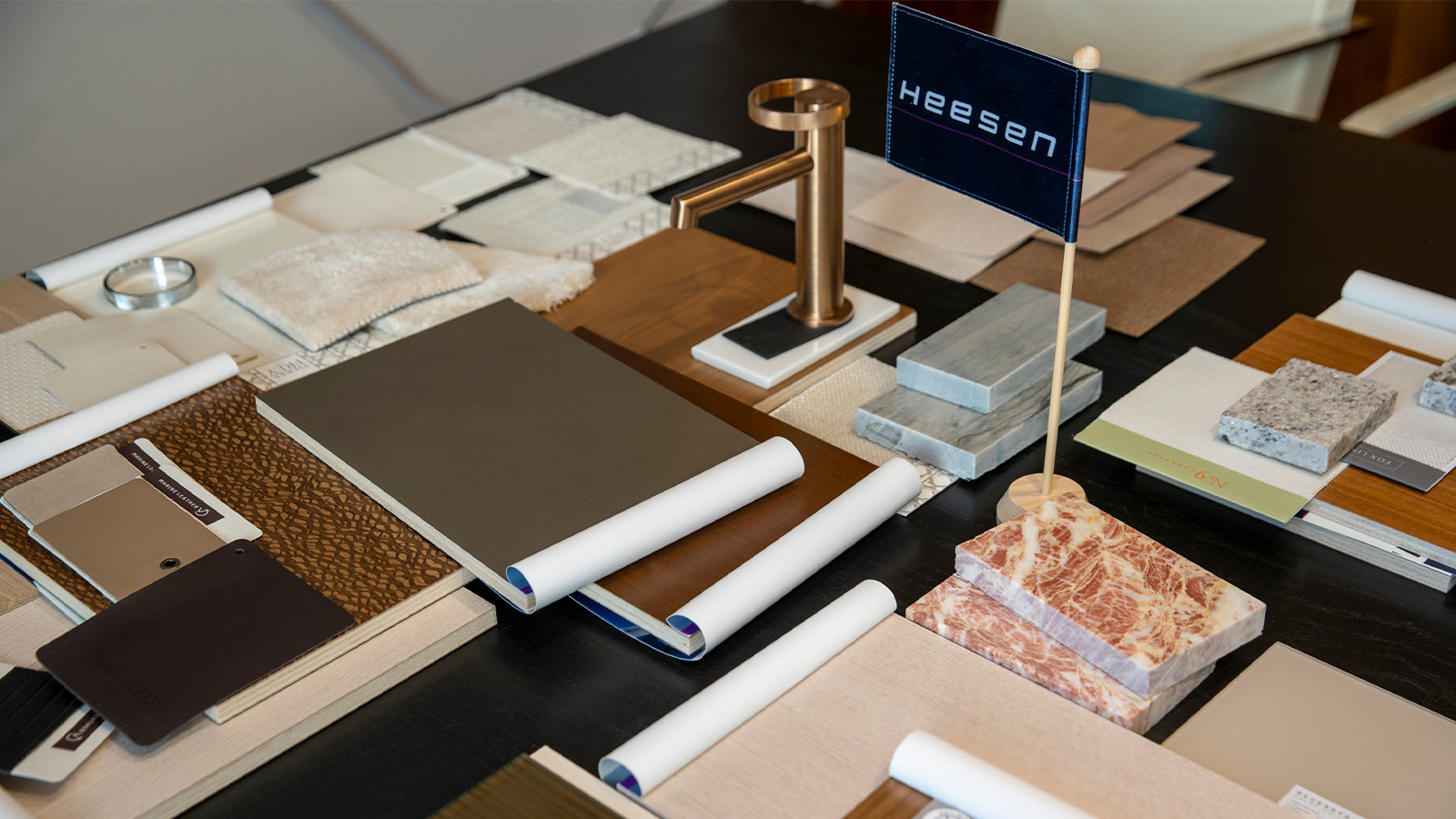 A collection of material samples including fabrics, wood, stone, and metal finishes are arranged on a table, featuring a small flag with "HEESEN" printed on it.