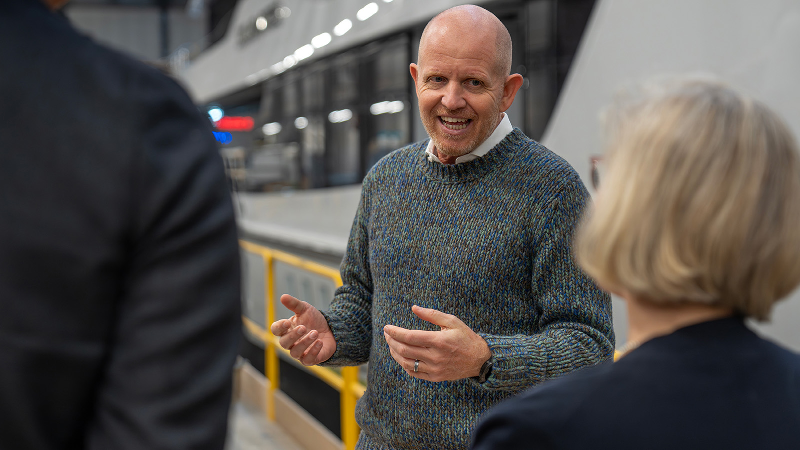 A man in a blue patterned sweater is speaking enthusiastically to two people in front of a large industrial or factory setting.