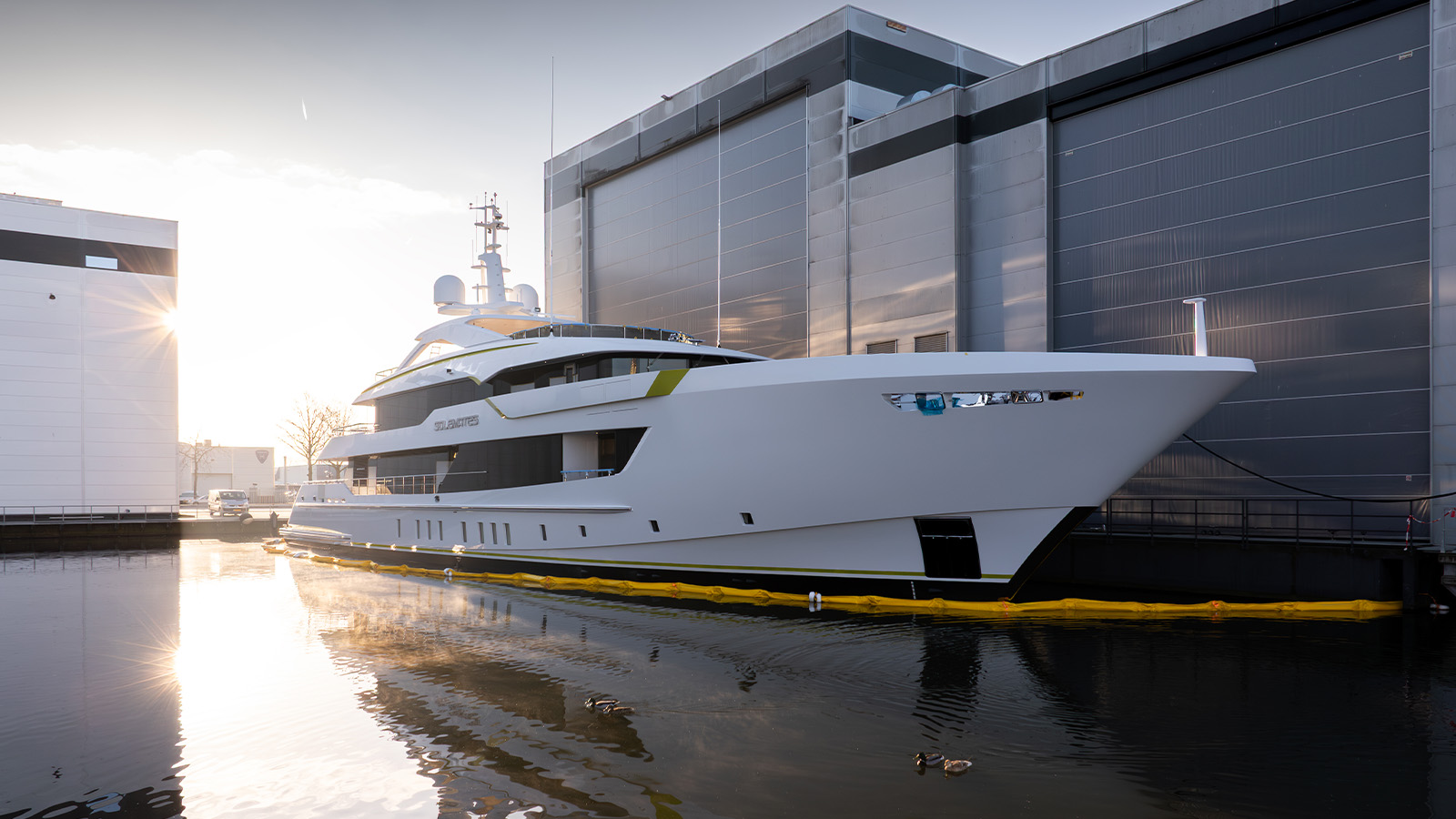 A modern luxury yacht docked at a shipyard, with the sun shining through buildings in the background and its reflection visible in the water.