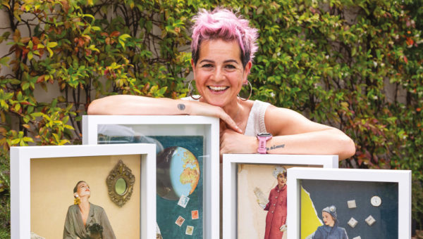 A person with short pink hair smiling and leaning on a set of framed collage artworks outside, with green foliage in the background.