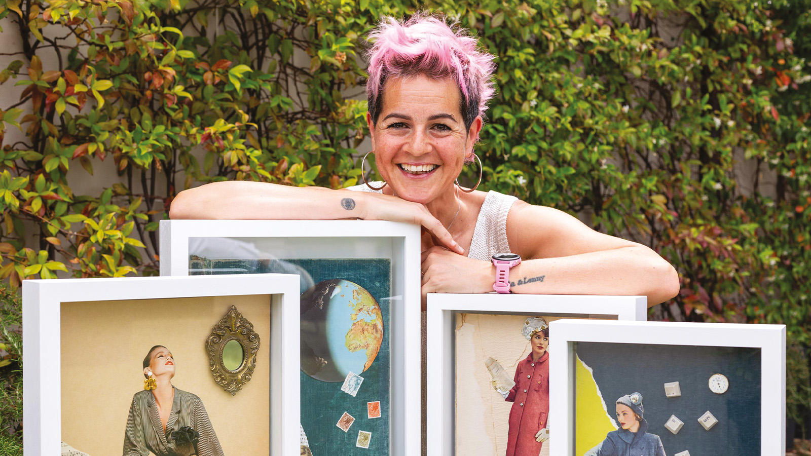 A person with short pink hair smiling and leaning on a set of framed collage artworks outside, with green foliage in the background.