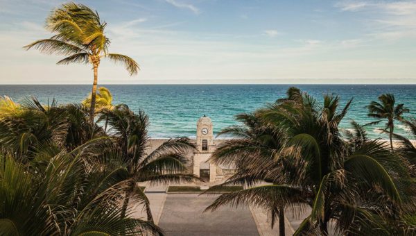 View of a tropical beach with swaying palm trees framing a clock tower structure near the shore against a blue ocean background.