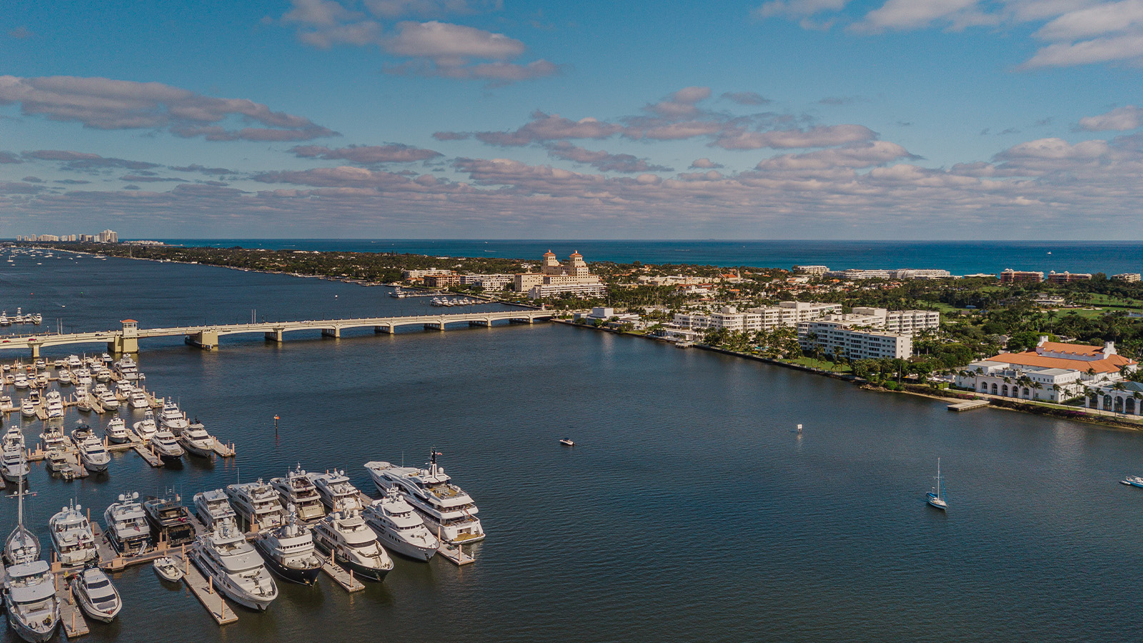 A coastal cityscape featuring a marina filled with numerous yachts and boats, a bridge spanning the water, and a mix of residential and commercial buildings along the shoreline. The sea stretches out into the distance under a partly cloudy sky.