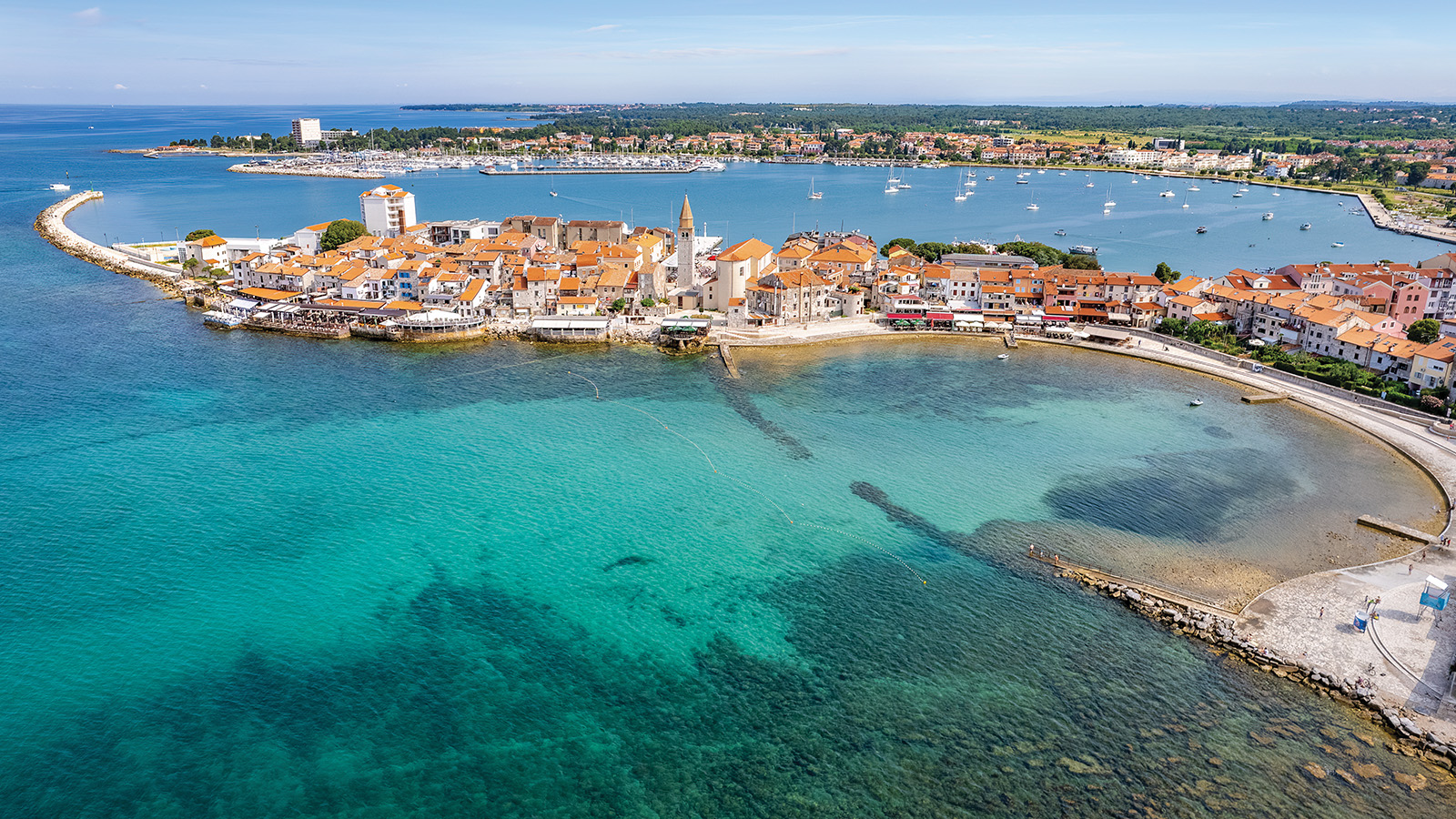 Aerial view of a coastal town with red-roofed buildings surrounded by clear blue waters, featuring a curved stone pier and several boats anchored nearby.