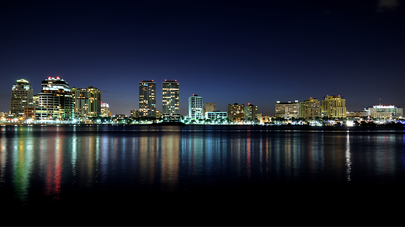 City skyline at night with illuminated buildings reflecting on the calm water below, against a dark blue sky.