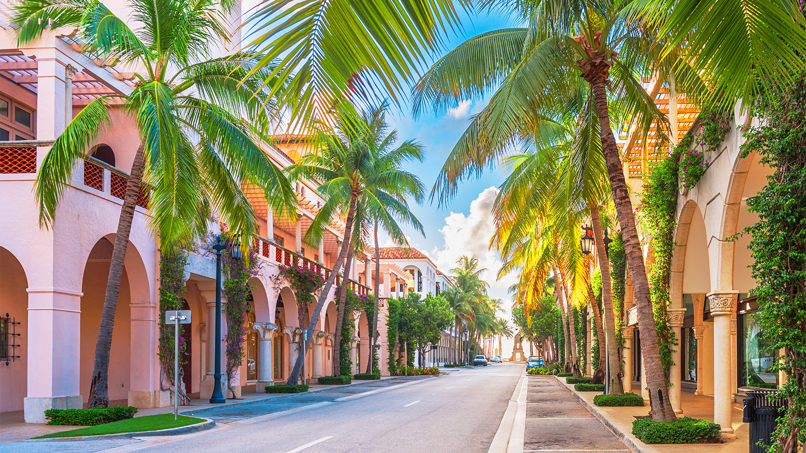 A scenic street in Palm Beach, Florida, lined with tall palm trees and buildings featuring Mediterranean-style architecture with archways.